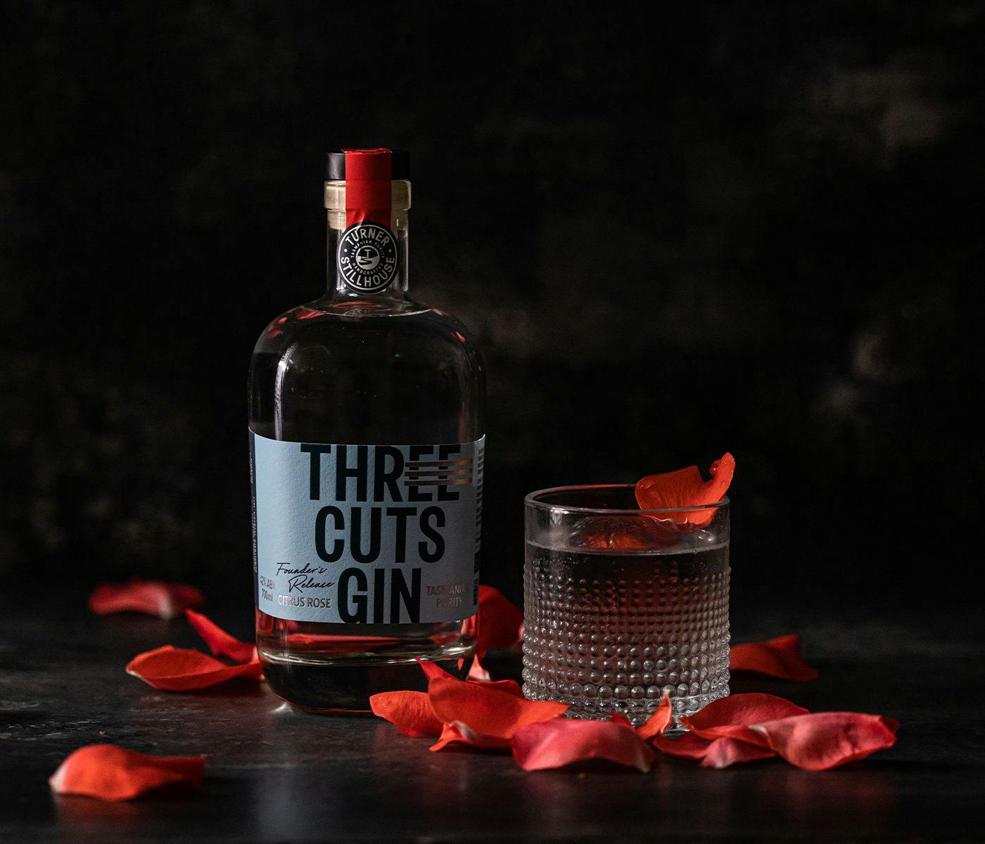 A bottle of Three Cuts Gin accompanied by a glass of gin and tonic, rose petals surround both.