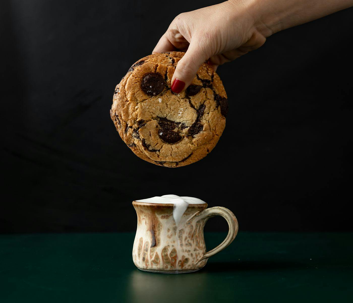 A rustic cup filled with frothy milk, a huge cookie (too big to dunk) is held above it.