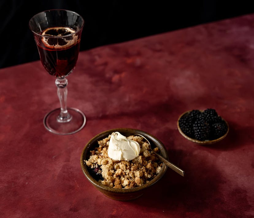 A bowl of apple and blackberry crumble, accompanied by a glass of red liquid and small bowl of blackberries.