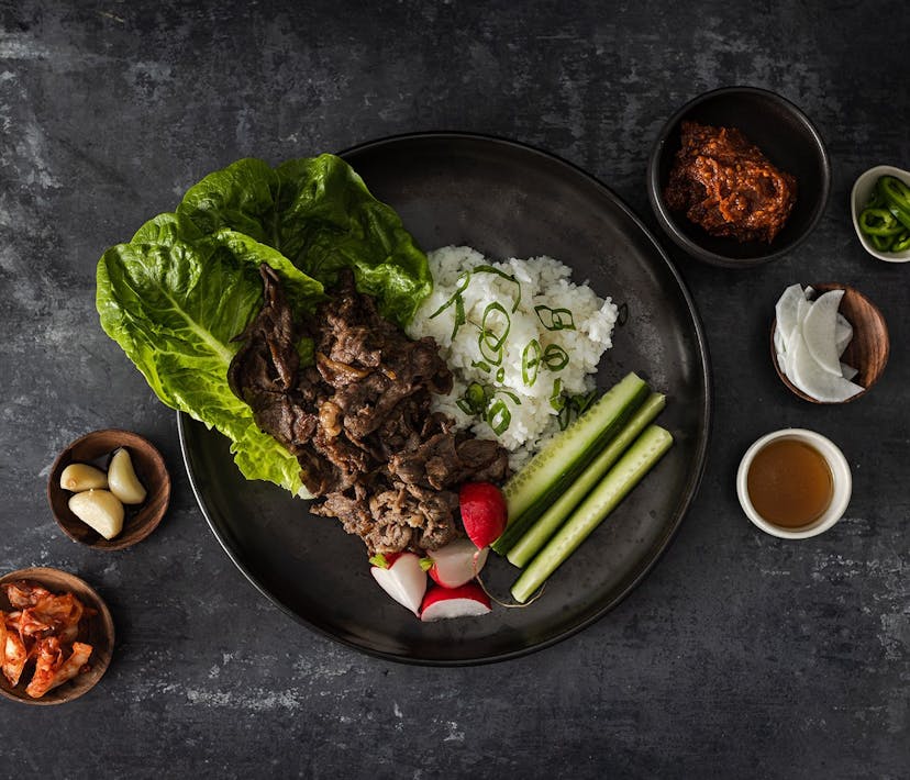 A plate of Bulgogi Ssam - rice, lettuce and meat, surrounded by various herbs and spices.