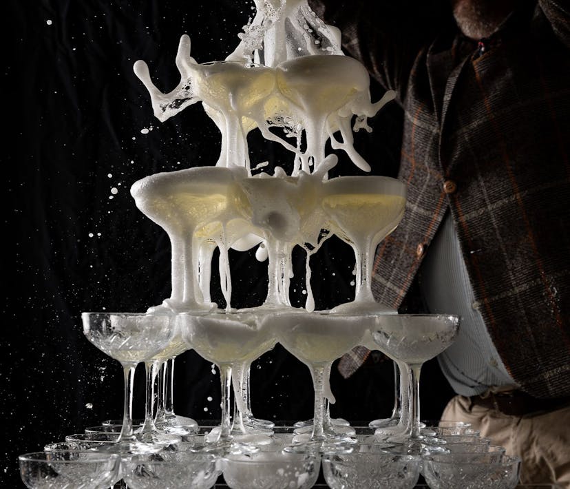 A pyramid tower of stacked cocktail glasses, sparkling wine is poured into the top layer, explosively flowing out of the glasses.