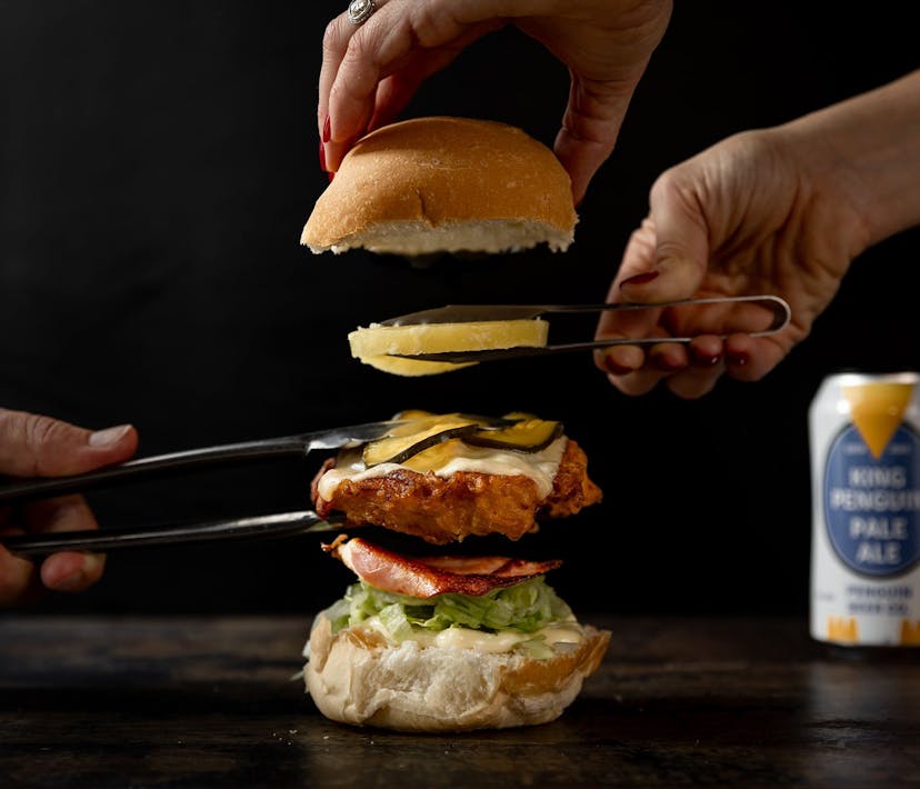 Three hands using tongs construct a stacked burger.