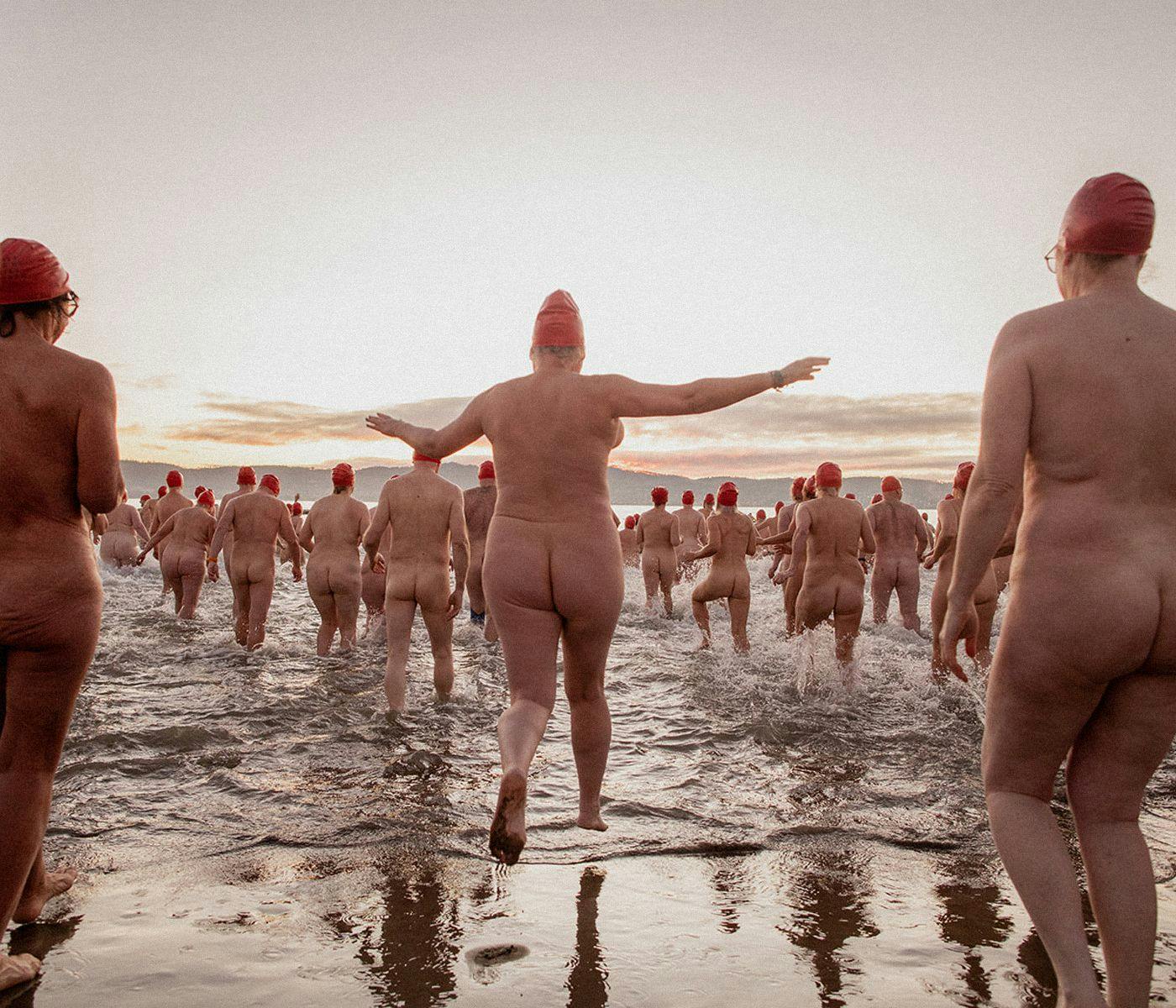 Several naked people wearing red swim caps run and jump into the ocean.