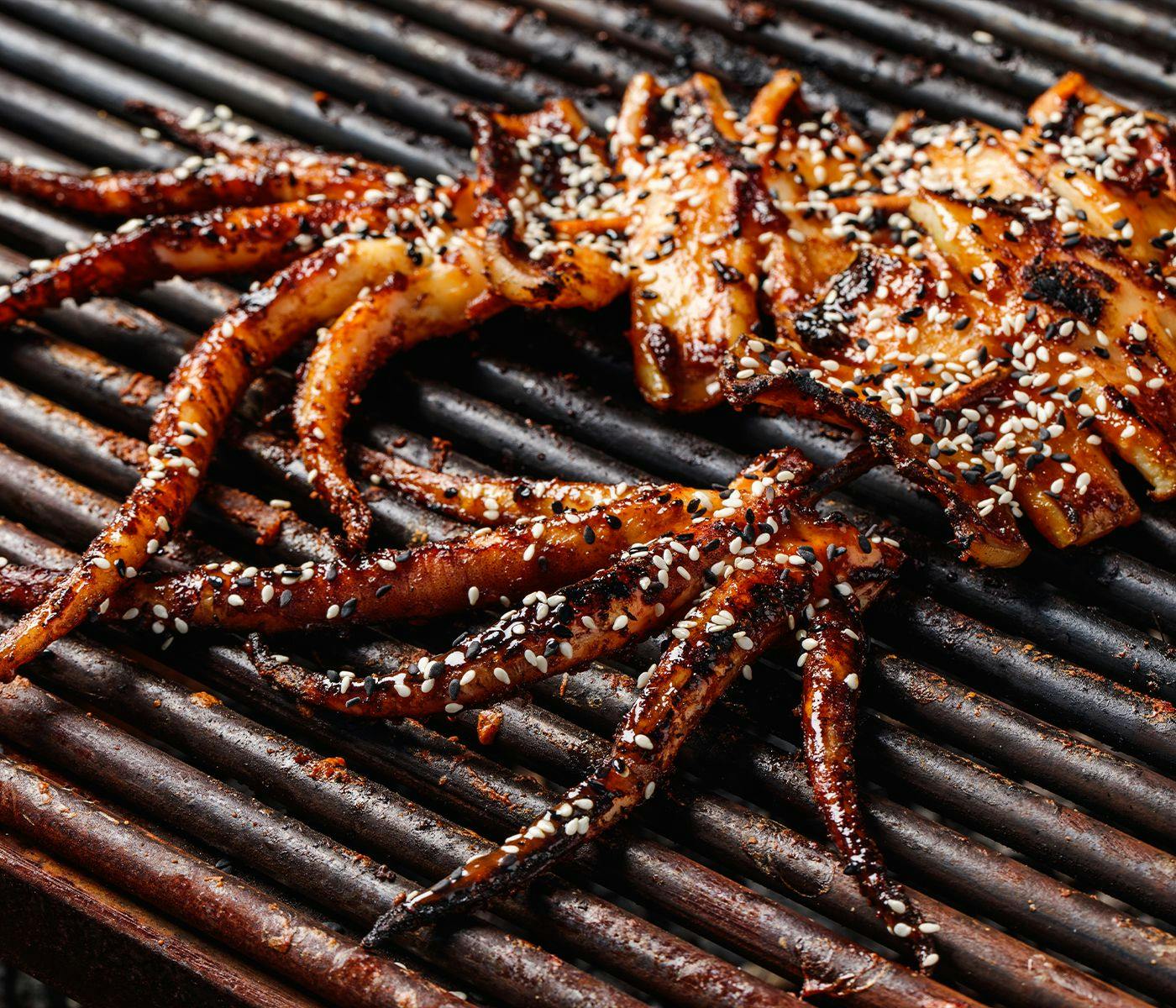 A large piece of charred squid, covered in sauce and sesame seeds, on top of grill.