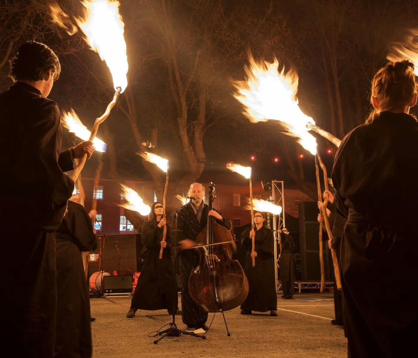 A person plays a cello, surrounding them are several black-robed people holding flaming sticks.