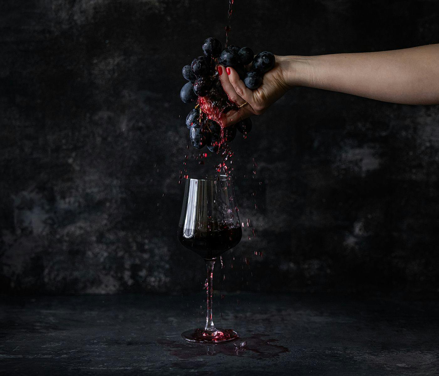 A hand crushes grapes, their juices flowing into a wine glass below.