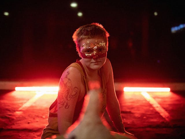A figure sits crouched on concrete wearing a sparkly mask, red lights light up the darkness behind them.