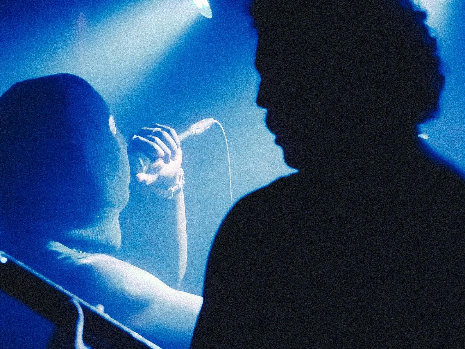 A silhouetted guitar player foregrounds a balaclava wearing person performing with a microphone.
