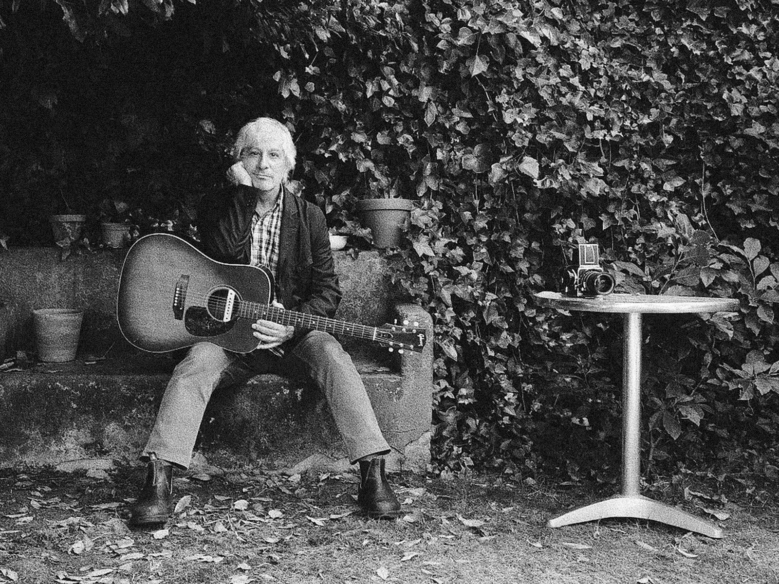 Lee Ranaldo sitting with his guitar on a stone bench.