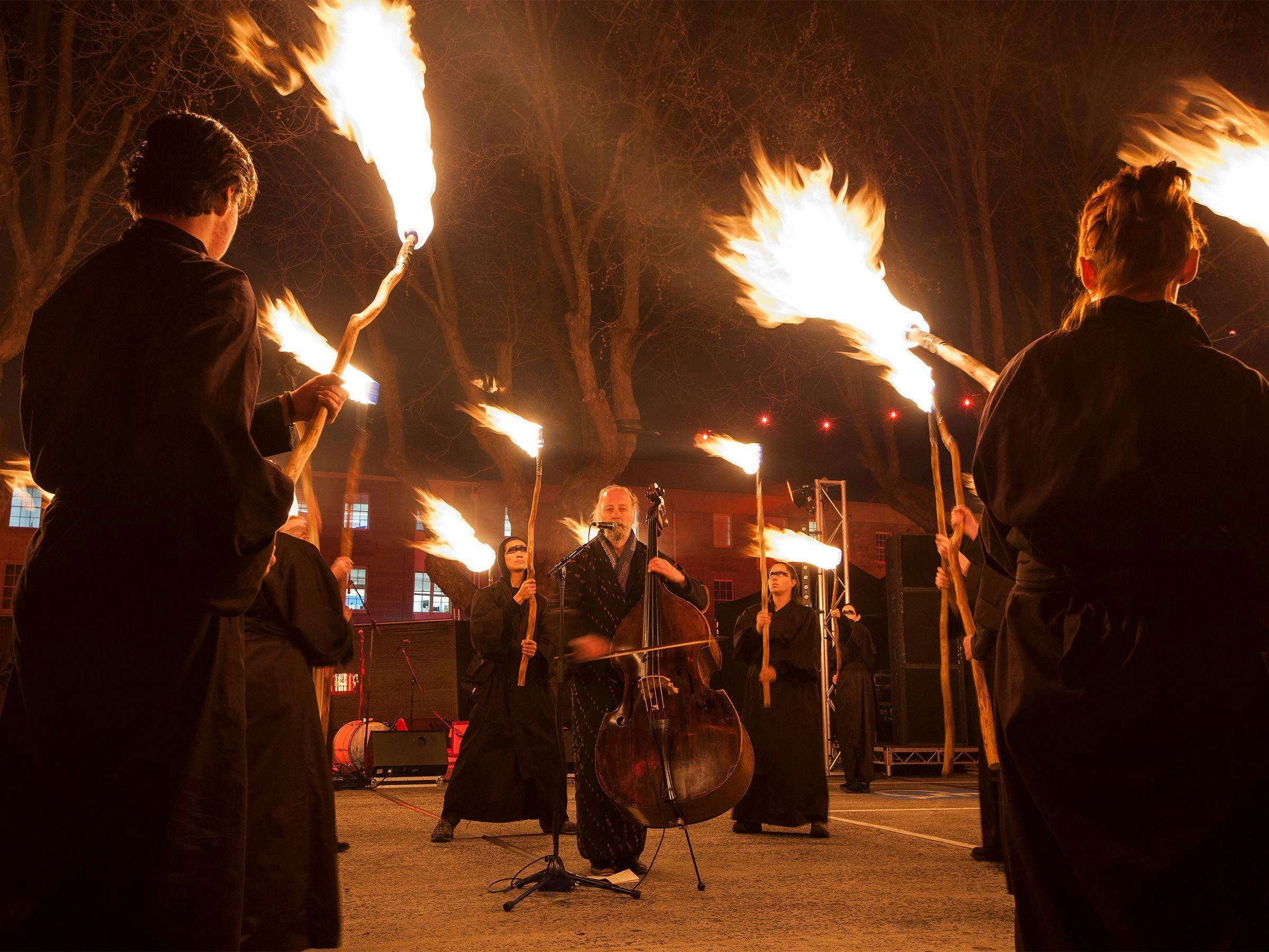 A man in a cloak playing the cello, surrounded by people holding fire sticks. 