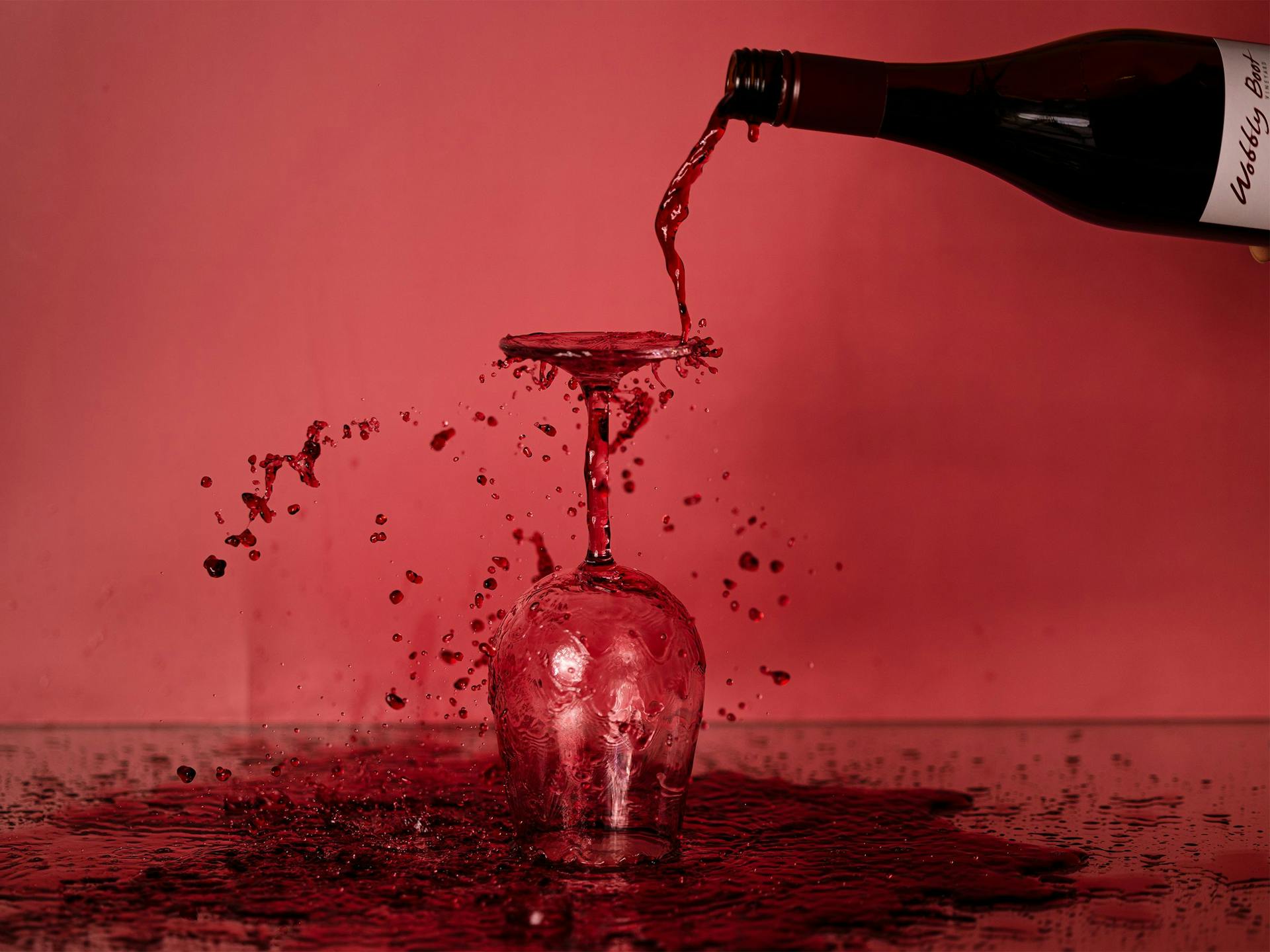 A bottle of red wine being poured on an overturned wine glass causing a big puddle