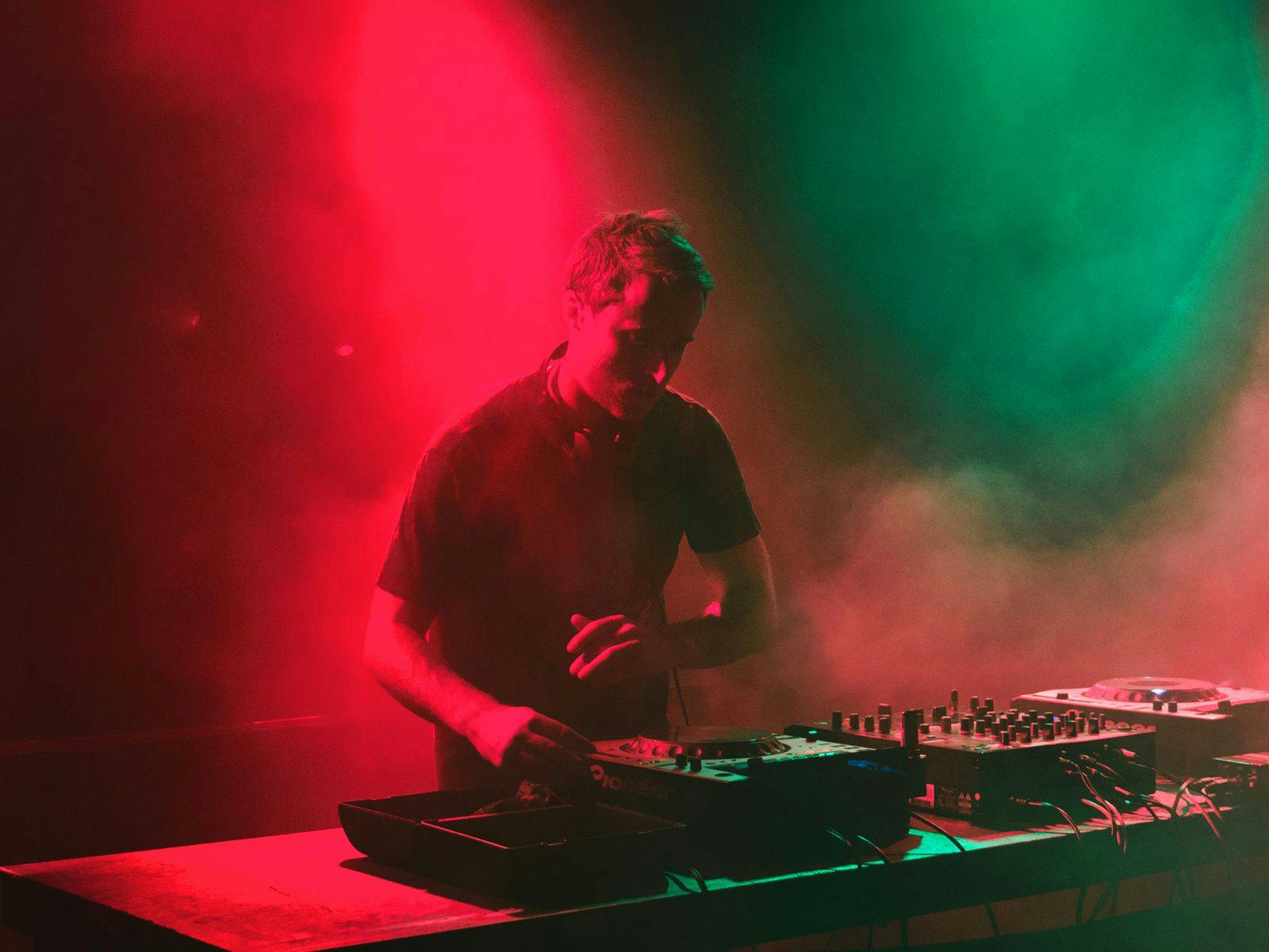 JLaw performing on a set of DJ decks, smoke fills the room with red and green lighting up the scene.