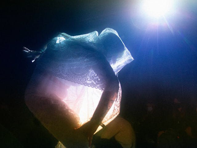 A lone figure wearing an ornate draped see through outfit. They are heavily backlit by a bright white and blue light.