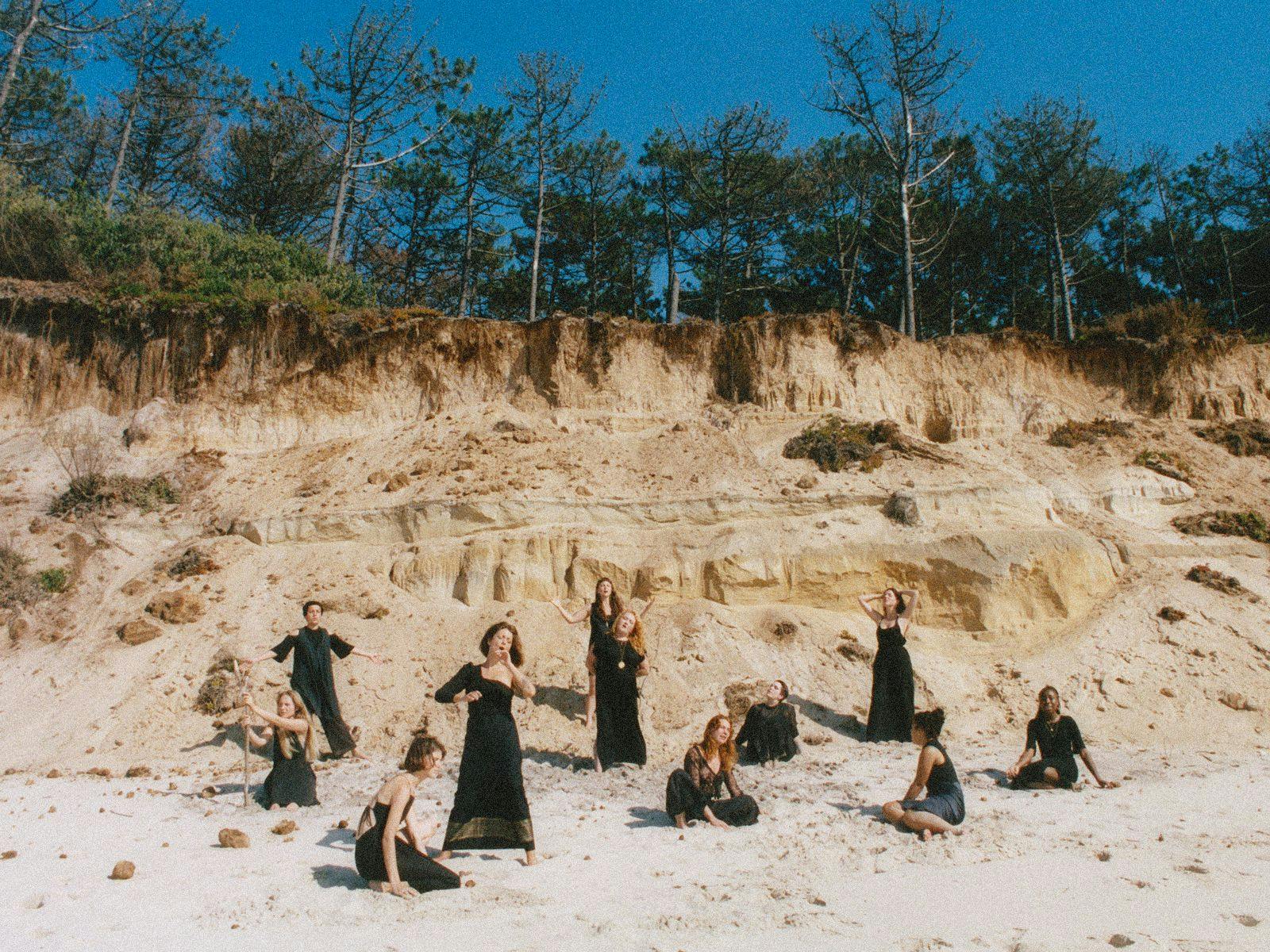 Several members of NYX pose on a beach, a large tree-topped crumbling sand dune backgrounds them.