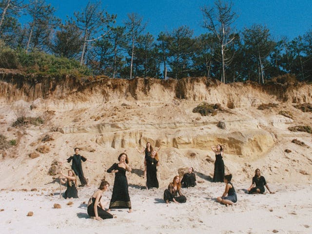 Several members of NYX pose on a beach, a large tree-topped crumbling sand dune backgrounds them.
