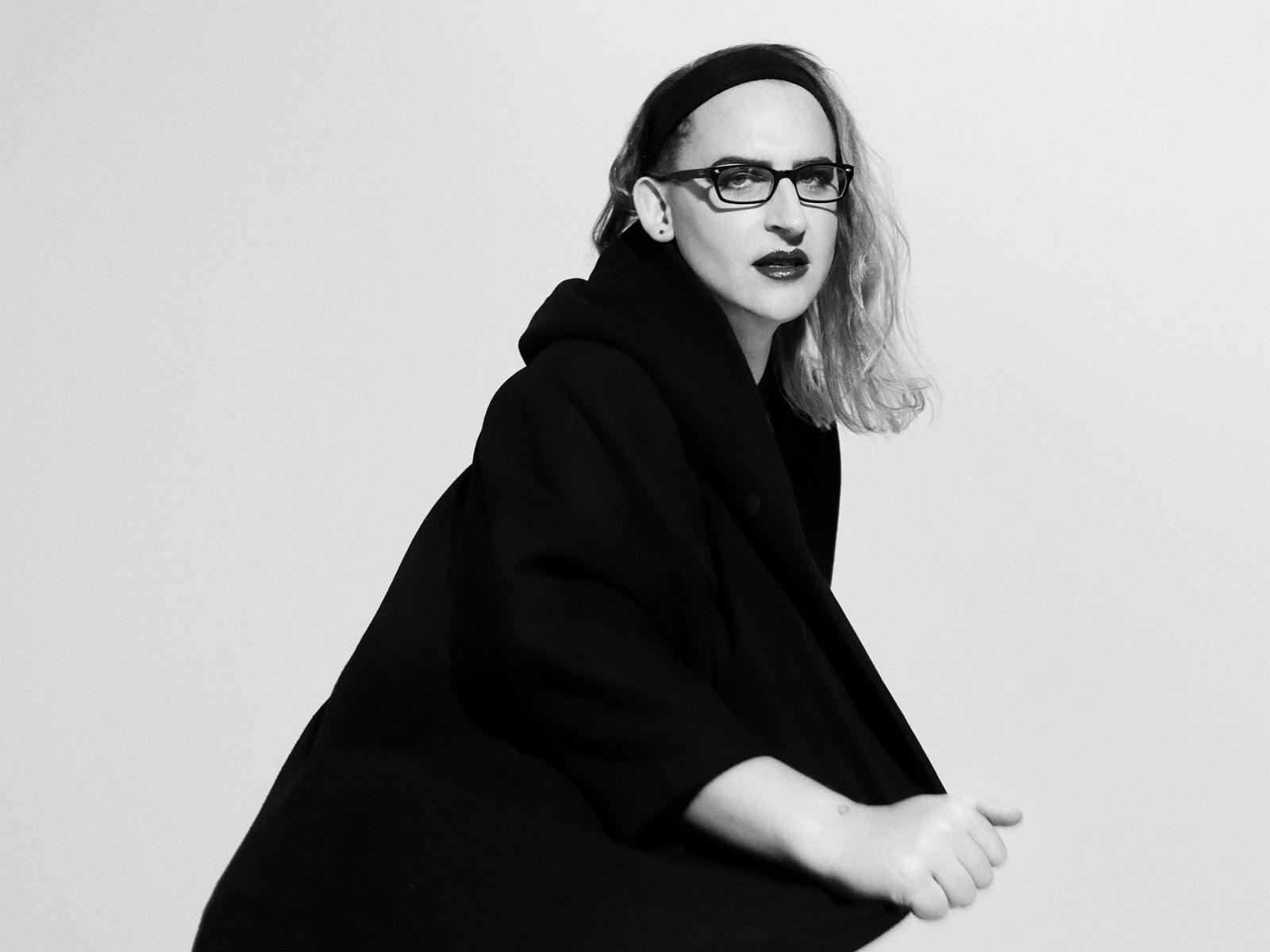 Simona Castricum crouches down, wearing a long black shawl, glasses and lipstick.