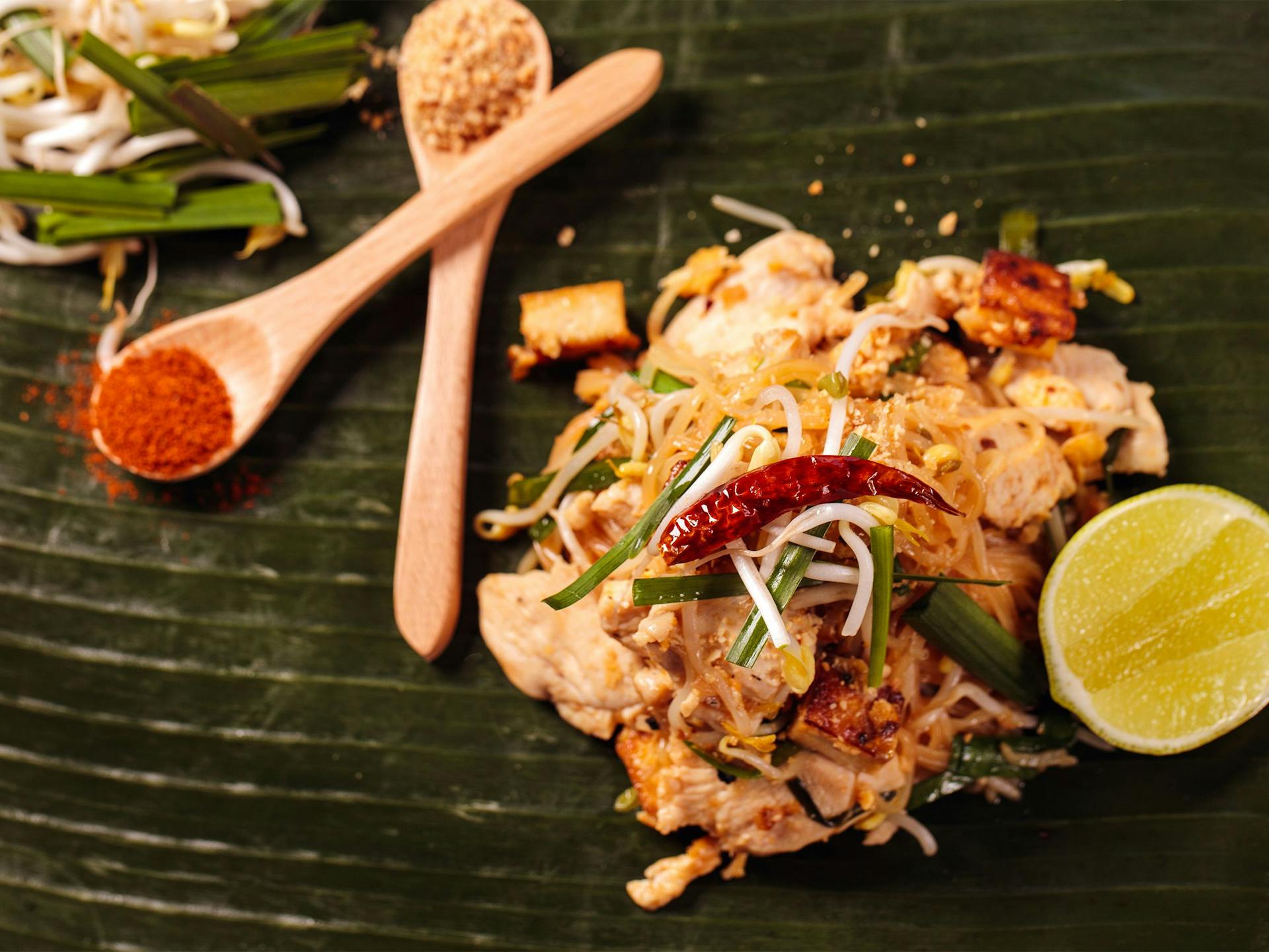 Pad Thai on a banana leaf, with spices in spoons and a lime wedge