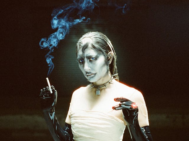 King Woman wearing heavily stylised makeup holds a burning cigarette in their hand.