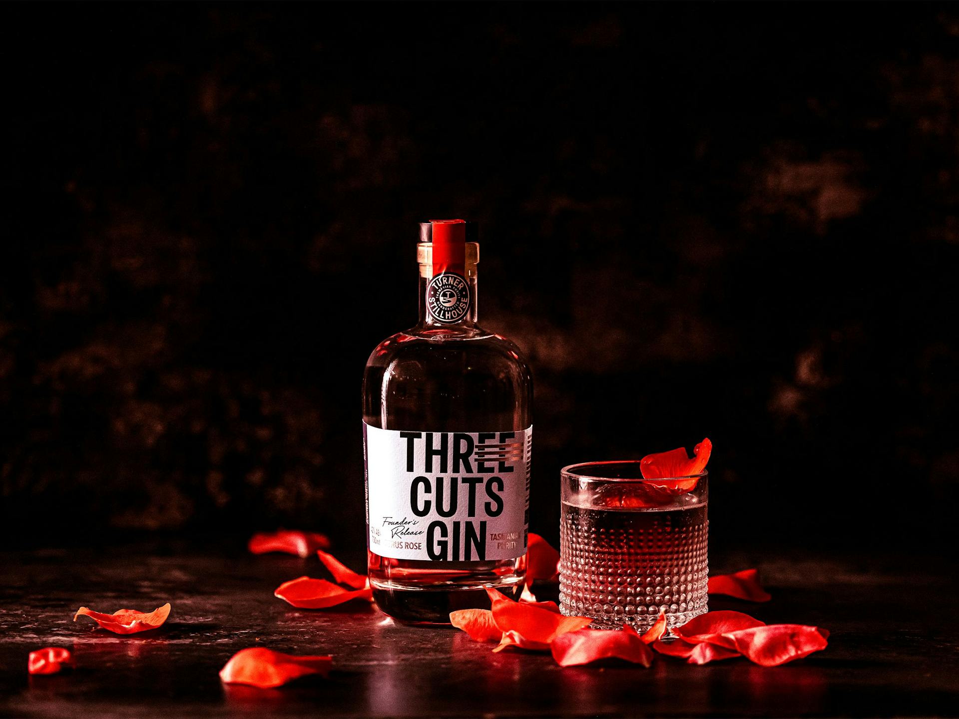 A glass and bottle of Three Cuts Gin surrounded by red rose petals
