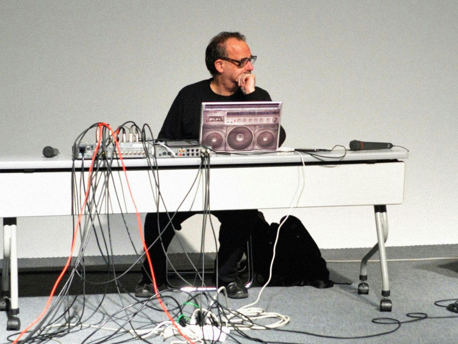 Carl Stone sits at a white desk, laptop, MIDI's and cables strewn around him.