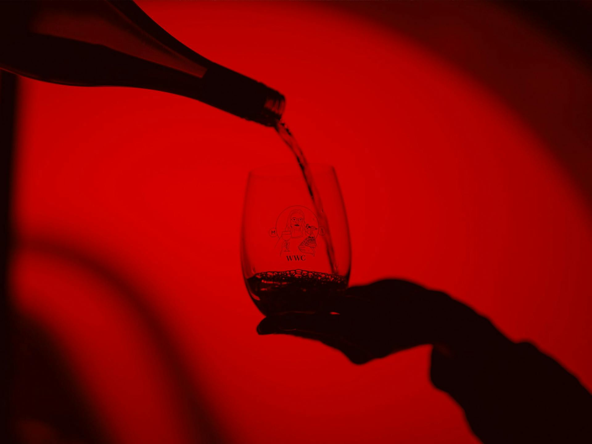 A glass of wine being poured against a bright red background. All you can see is the silhouette of the hand holding a glass and the shadow of the bottle pouring white wine.