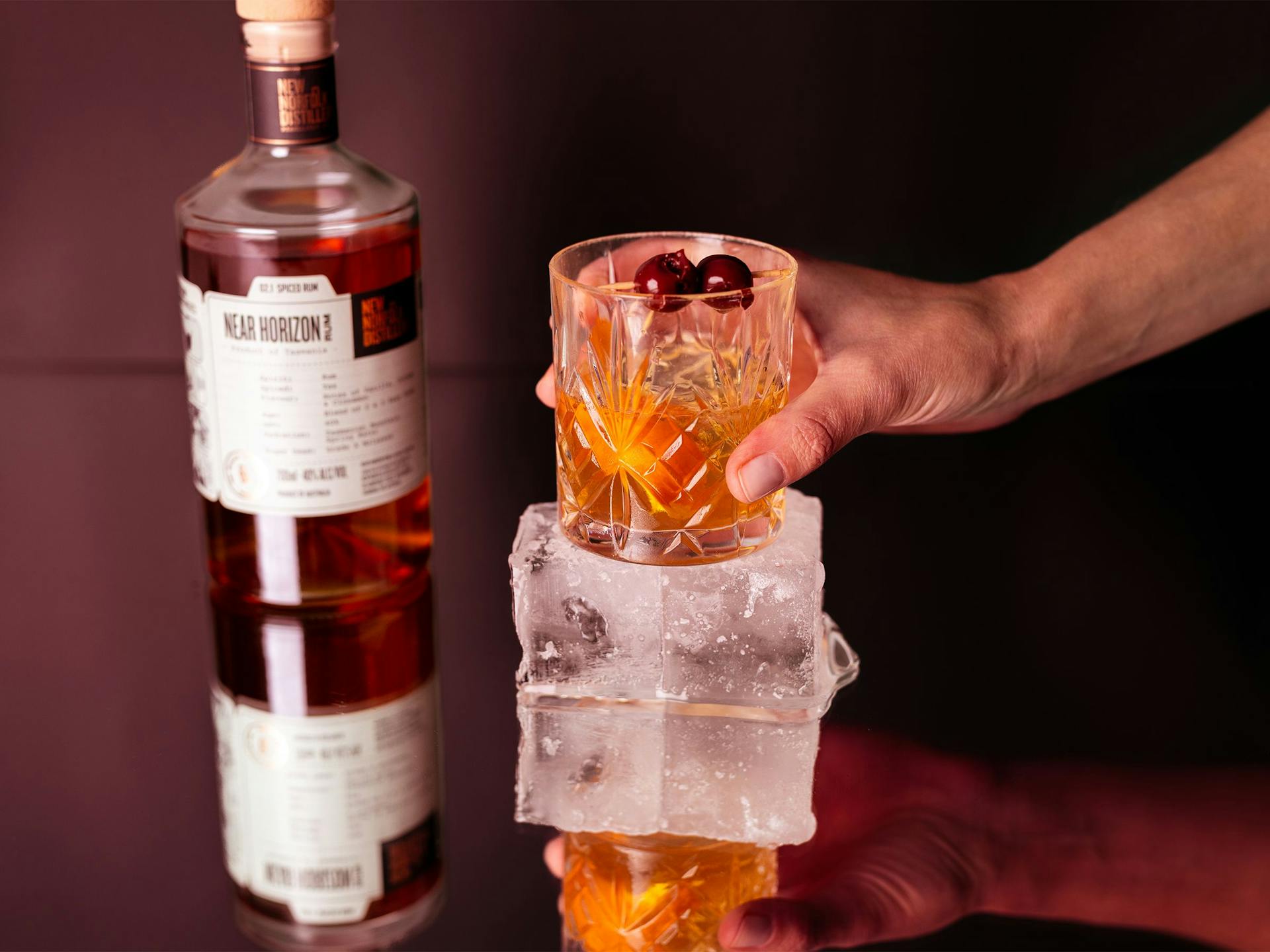 A bottle of Near Horizon rum sits behind a glass resting atop a big ice cube. The whole scene is being reflected on a mirrored surface