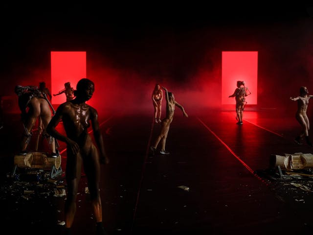 Several naked women perform various motions on a large stage, red lighting and smoke visible behind them.