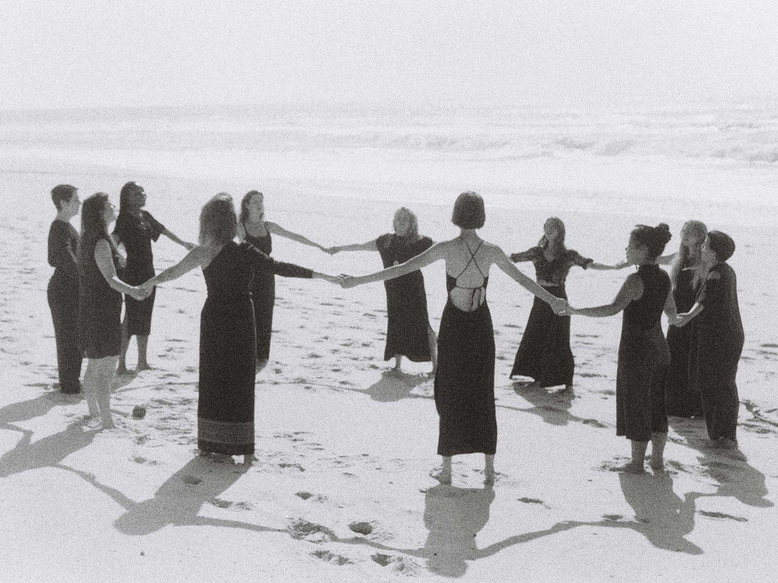 A group of 11 people stand in a large circle on a beach, their hands held to join the circle.