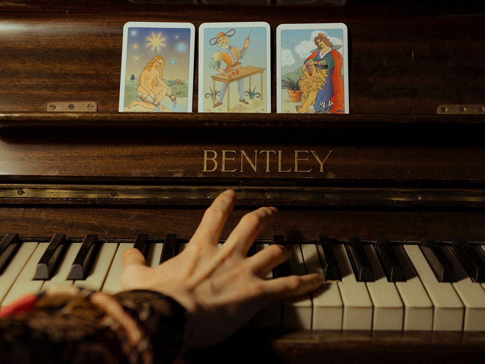 A close-up of a hand playing a Bentley piano. Three tarot-like cards sit on the music sheet sill.