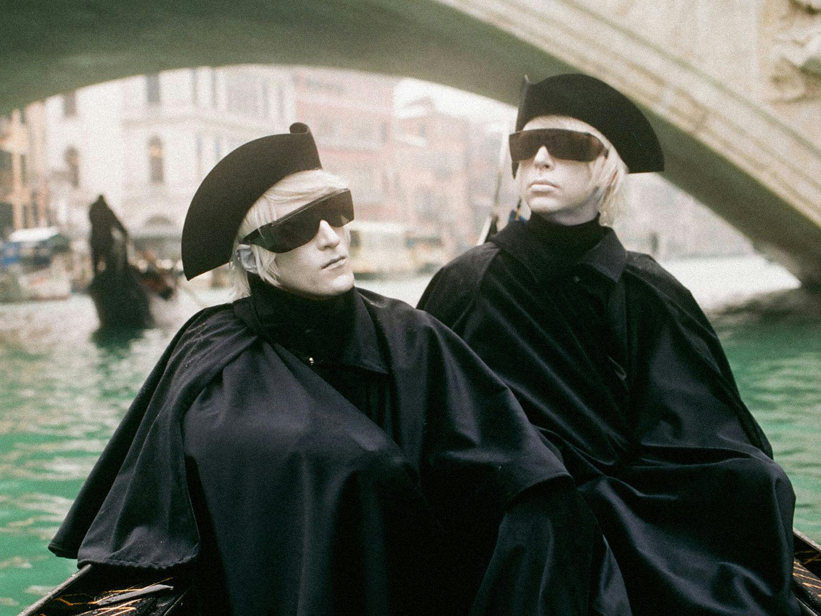 Members of Drab Majesty sit in a canal boat, wearing all black outfits, hats and sunglasses.