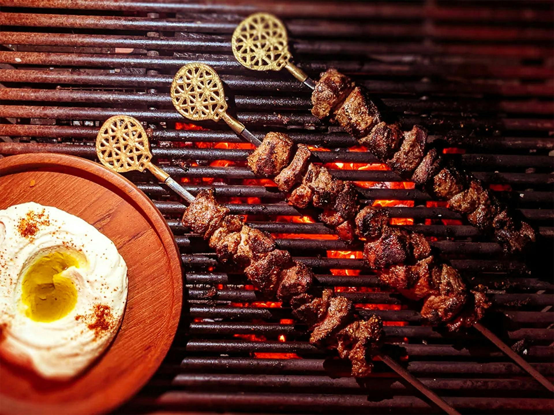 Three lamb kebabs on the grill with a side of hummus