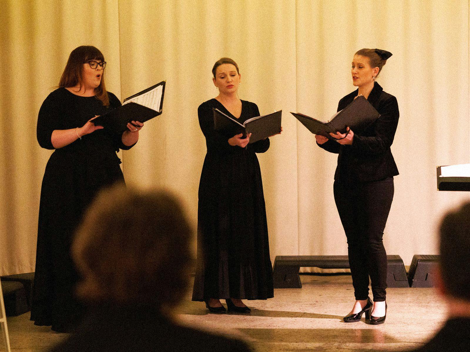 3 singers perform against a curtained background, dressed in fully black outfits they hold black folders of music notation.