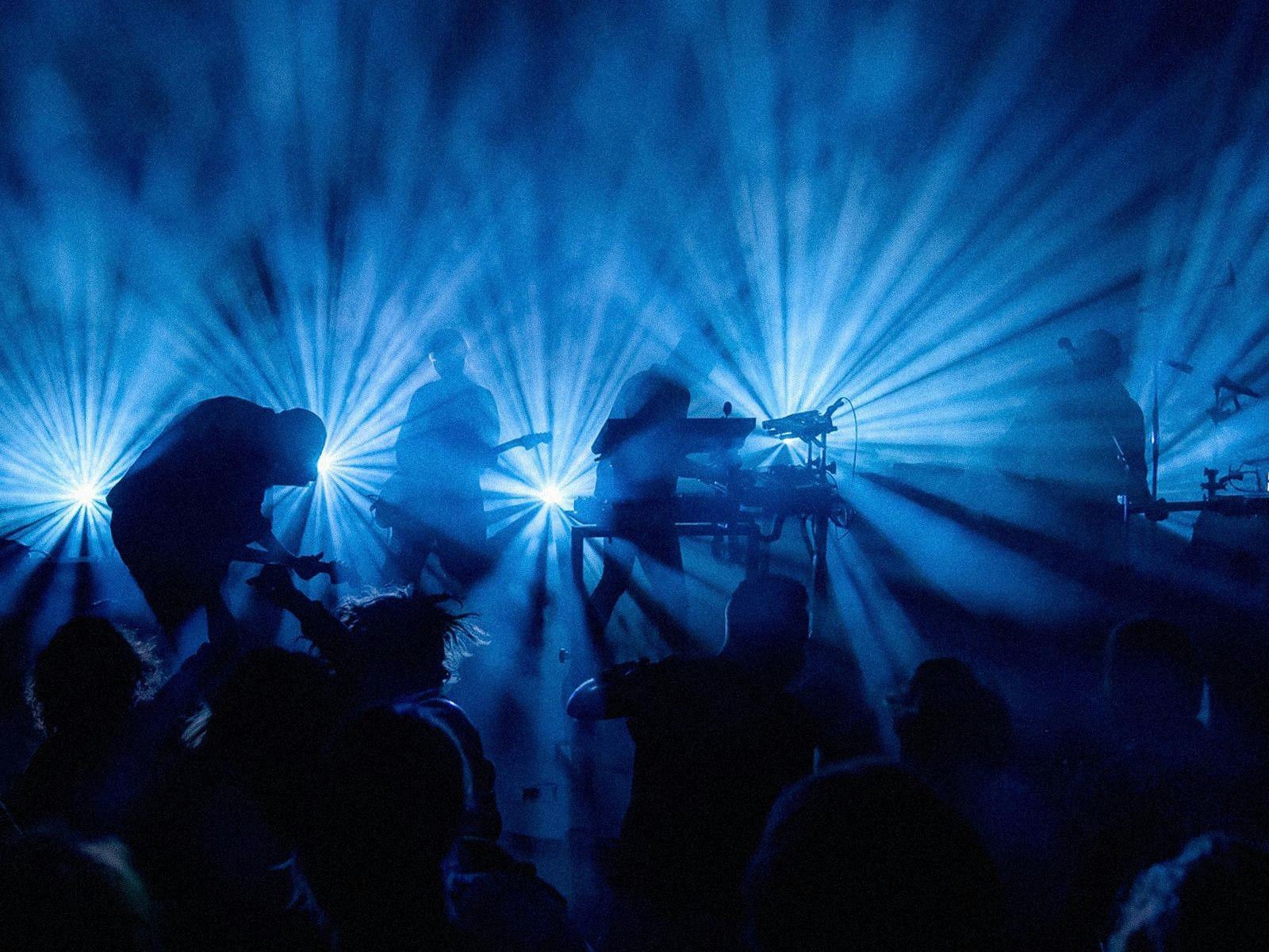 Silhouetted by dazzling blue lights, Trentemøller performs on stage with guitars and keyboards.