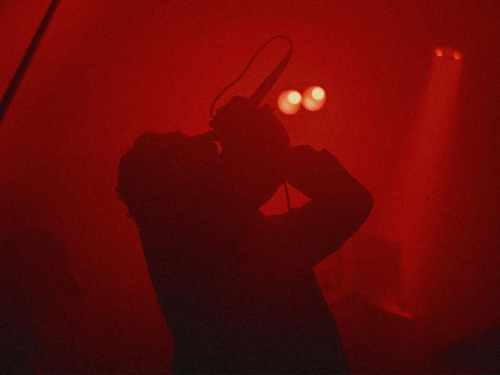 Member of Drowning Horse sings into a microphone. Around them is a heavily hazed stage, lit by red lights.